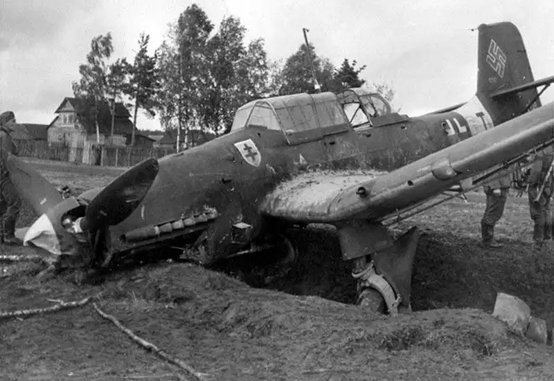 Black and white historical photo of a damaged German Junkers Ju 87B Stuka dive bomber resting in a ditch near a Soviet wooden village house in 1942.