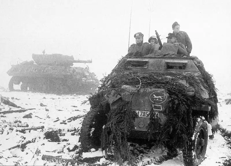Black and white WWII photo of a German camouflaged Sd.Kfz. 251 half-track in snow with German soldiers on board and a destroyed US M10 Wolverine tank destroyer in the background near Saint-Vith, December 1944.