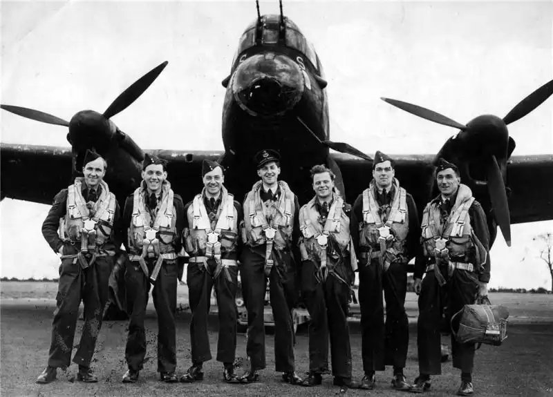 Seven RAF aircrew members posing lined up in front of an Avro Lancaster bomber nose section, 1943.