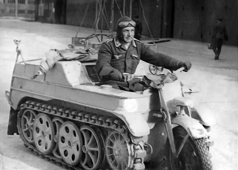 Luftwaffe serviceman wearing a leather flight helmet driving a light half-track NSU Kettenkrad Sd.Kfz. 2 on an airfield in 1944.