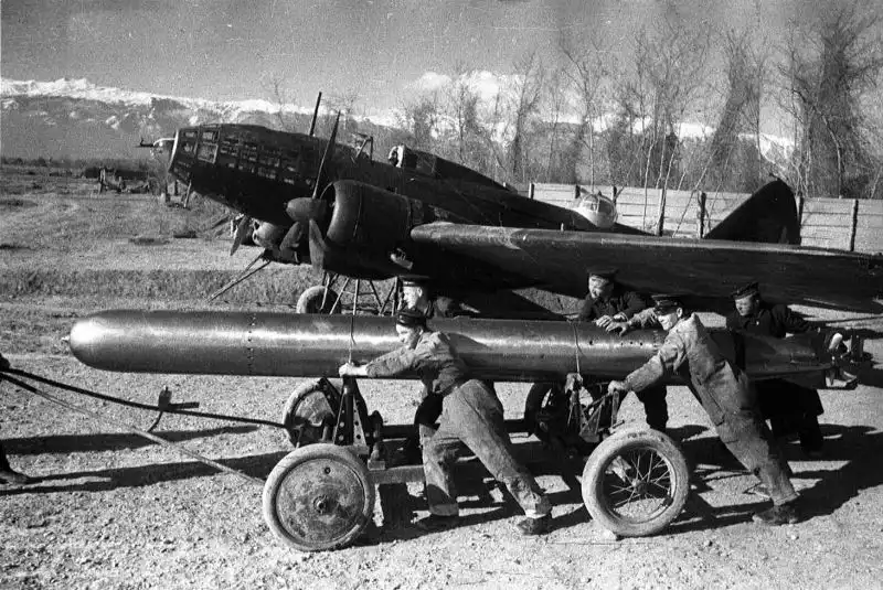 Soviet naval armorers pushing a long 45-36AN aerial torpedo on a cart toward an Il-4T torpedo bomber at Gudauta airfield with snow-capped mountains in the background in 1942.