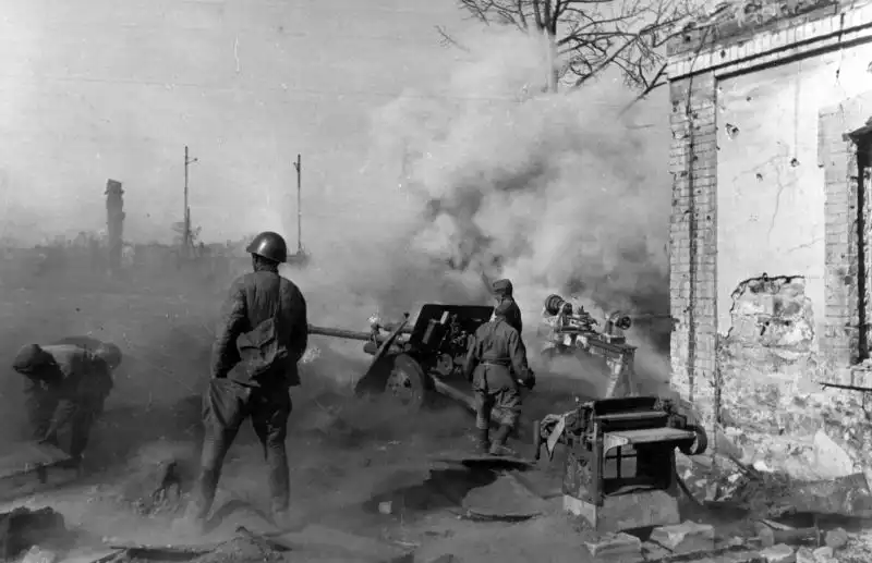 Black and white historical photo by Arkady Shaikhet showing a Soviet 76mm ZiS-3 anti-tank gun firing near factory machinery in ruined Stalingrad, 1942.