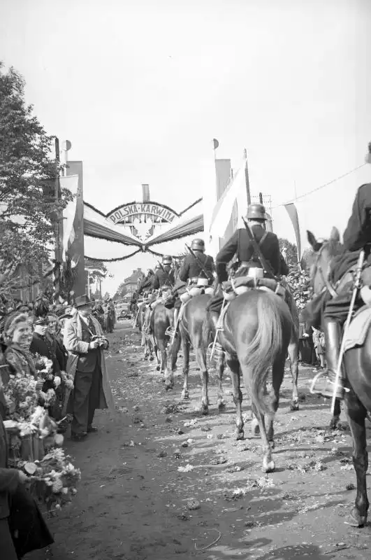 Black and white historical photo from NAC showing Polish cavalry soldiers riding past crowds and a decorated archway reading "Polska-Karwina" in October 1938.
