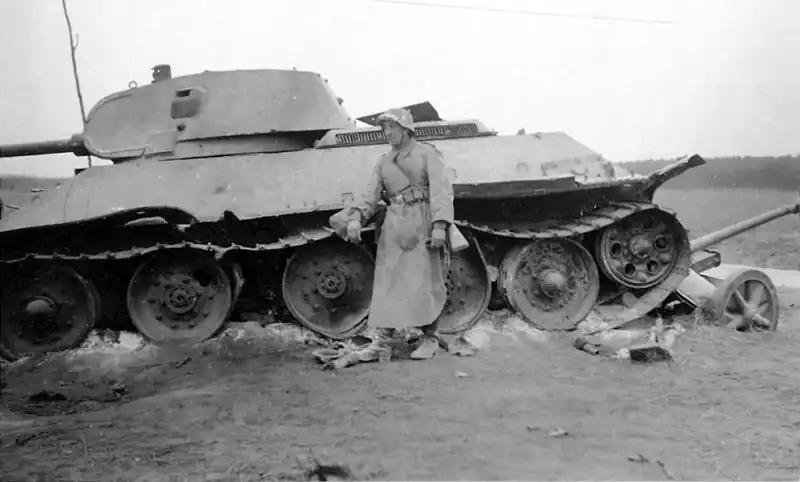 Black and white historical photo of a damaged early Soviet T-34 tank resting on top of a crushed German 5cm PaK 38 anti-tank gun with a German soldier nearby, June 1941.