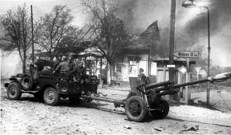 Red Army artillerymen riding in a Lend-Lease Dodge WC-51 truck towing a 76.2mm ZiS-3 gun past a burning building in Germany, April 1945.