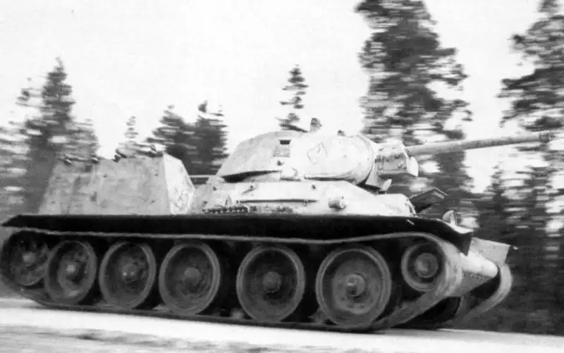 Soviet T-34 medium tank in white winter camouflage moving along a forest road, fitted with armored covers for FOG-1 flamethrowers on its rear deck in 1942.
