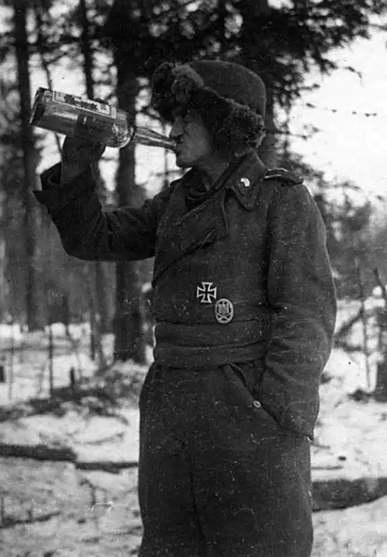Black and white historical photo of a German self-propelled gun crewman wearing a field-grey jacket and winter hat holding a bottle on the Eastern Front in 1944.
