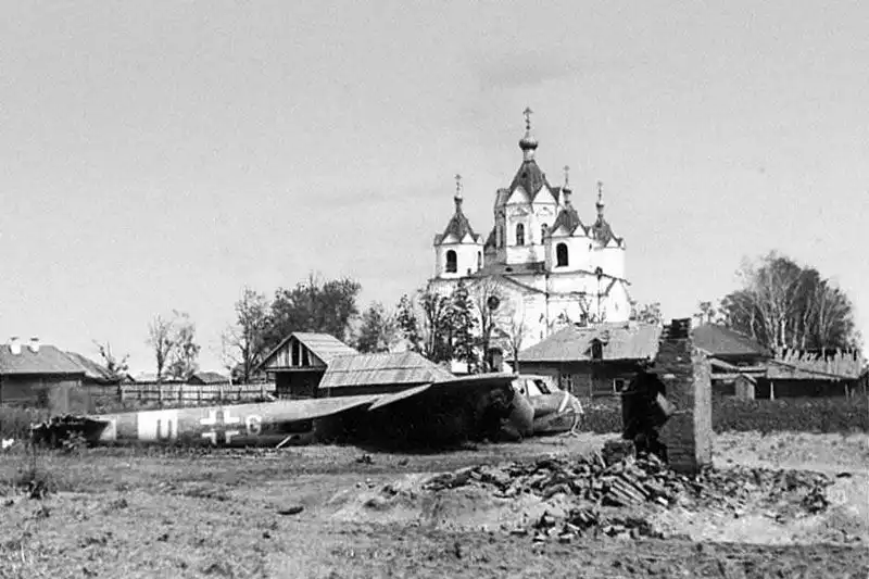 Black and white WWII photo of a crashed German Dornier Do 17Z bomber lying on the ground with an old Russian Orthodox church in the background in Demidov, 1941.