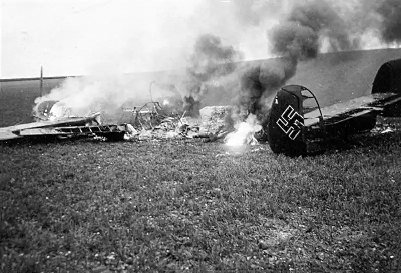 Black and white historical photo of the tail section of a burning German Messerschmitt Bf 110 twin-engine fighter lying in a field in Poland in 1939.