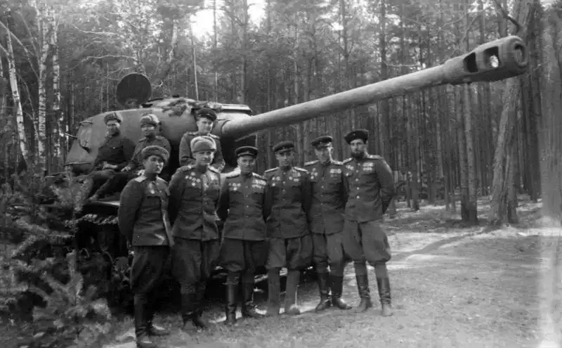 Black and white historical photo showing Soviet Red Army officers standing in front of a massive IS-2 heavy tank in a pine forest, April 1945.