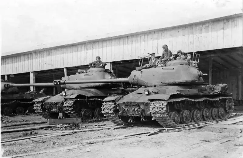 Black and white historical photo showing Soviet IS-2 heavy tanks with crew members lined up in a covered wooden tank hangar in Güstrow, July 1945.