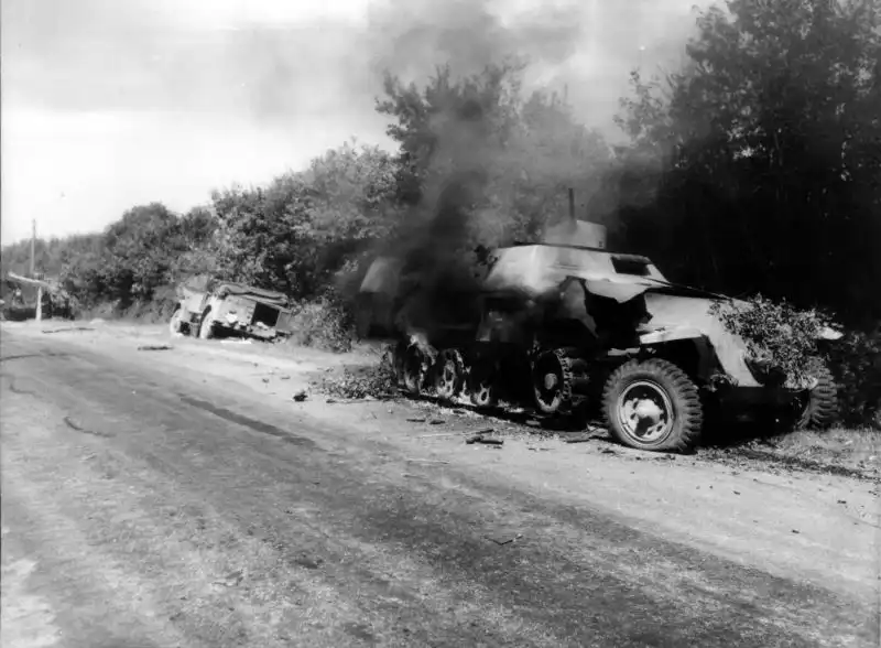 Black and white historical photo of a burning German Sd.Kfz. 251 half-track and a destroyed truck on a French country road in Normandy, August 1944.