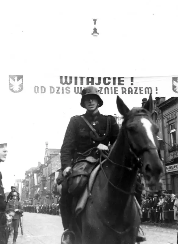 Black and white historical photo from NAC showing a Polish mounted policeman in a helmet riding through a street decorated with a welcoming banner in Český Těšín, 1938.