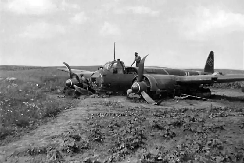 A German Junkers Ju 88 bomber resting on its belly in a plowed field with bent propeller blades as ground personnel inspect the aircraft in 1942.