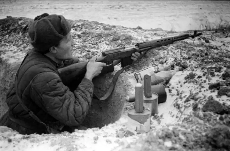 Black and white archival photo of a Soviet soldier in a trench holding an SVT-40 semi-automatic rifle with anti-tank grenades on the parapet.