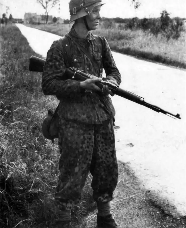 Young German SS grenadier standing on a rural road in Normandy wearing dot-pattern camouflage uniform and holding a Mauser K98k rifle in June 1944.