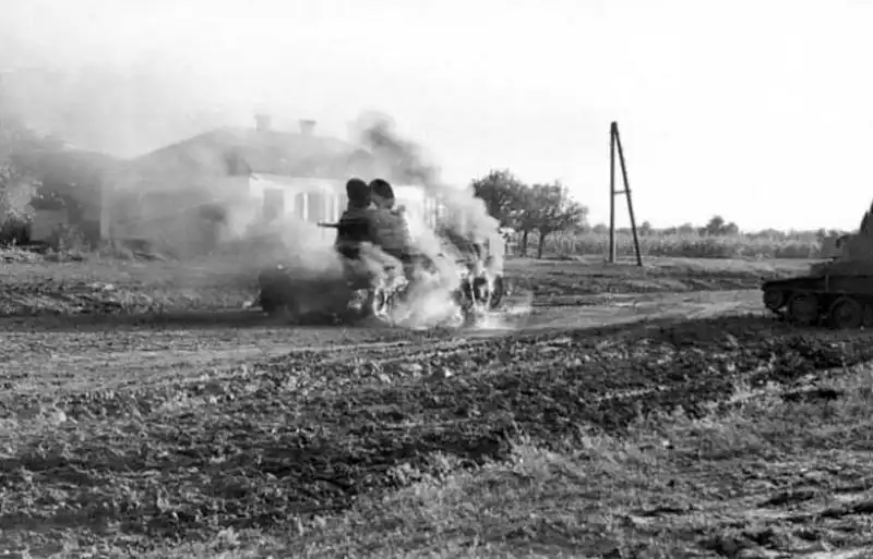Burning Soviet BT-7 light tank with open turret hatches on a dirt road next to another disabled BT-7 in summer 1941.
