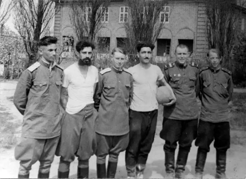 Black and white historical photo showing six Soviet Army officers and soldiers standing together outdoors holding a volleyball in Güstrow, July 1945.