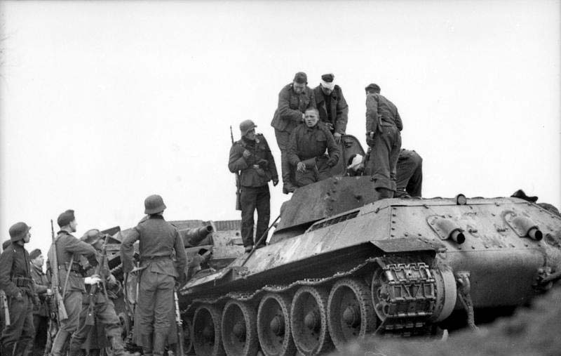 Black and white historical photo showing German soldiers surrounding a damaged T-34 tank and pulling out wounded Soviet tankers from an opened turret hatch in October 1941.
