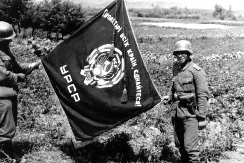 Black and white WWII photo of German soldiers displaying a captured Soviet Ukrainian SSR flag with Ukrainian inscription during Barbarossa in 1941.