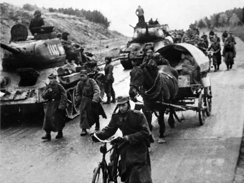 Black and white historical photo showing Soviet-supplied T-34-85 tanks with Polish markings moving along a paved highway alongside Polish infantrymen, cyclists, and horse carts in 1945.