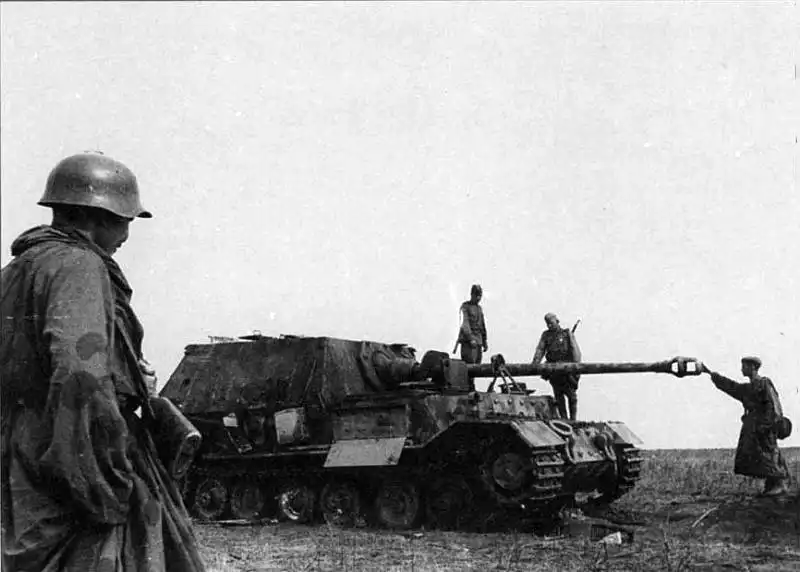 Black and white historical photo showing Soviet soldiers examining a large German Ferdinand tank destroyer in an open field, with a soldier in an SSh-36 helmet in the foreground, 1943.