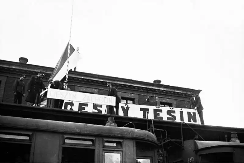 Black and white historical photo by Czesław Datka showing people on a station roof covering the large "Český Těšín" sign with a banner reading "Cieszyn Zachodni" in 1938.