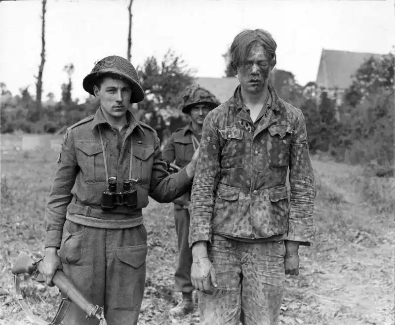 Black and white WWII photo of Canadian soldiers with Sten submachine guns standing with a bruised young German SS prisoner in Normandy in 1944.