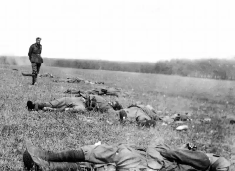 Line of fallen Soviet infantrymen in boots and puttees lying in an open summer field while a lone German soldier stands observing in the distance in 1941.