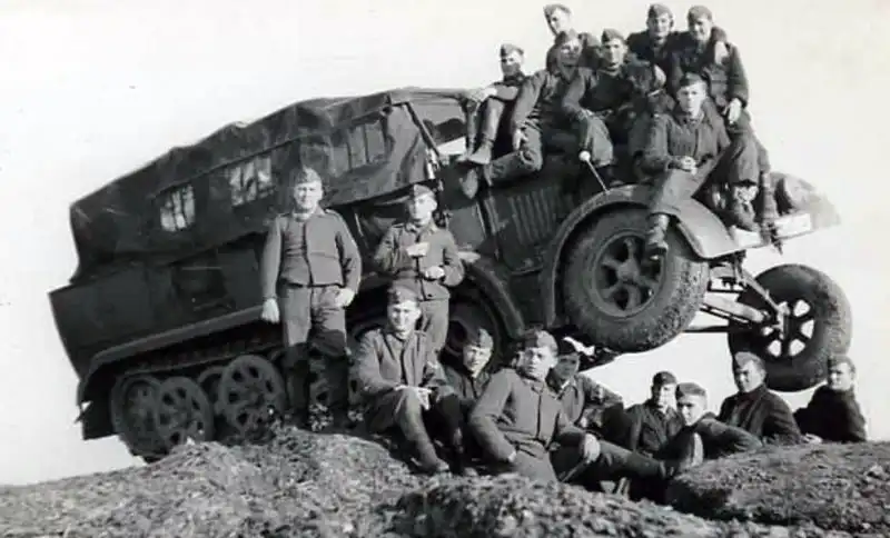A heavy German Sd.Kfz. 8 half-track tractor angled on a dirt mound with dozens of soldiers posing on the bonnet, roof, and side panels.