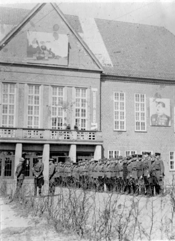 Black and white historical photo showing Soviet soldiers lined up in formation in front of a brick barracks building in Güstrow, July 1945.