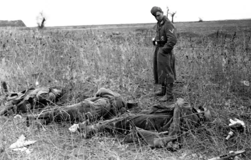 German Gefreiter in a winter coat standing over fallen Soviet soldiers laid out on a dry grass field alongside an unused RGD-33 grenade and a broken Kar98k rifle in autumn 1941.