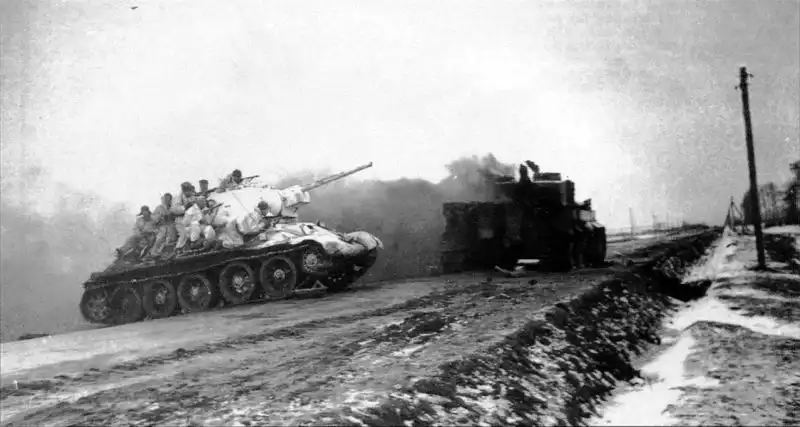 Black and white historical photo showing a Soviet T-34 medium tank carrying infantry in white winter uniforms crossing a road, with a burning German Tiger tank in the background, January 1944.