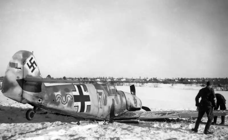 A German Messerschmitt Bf 109F-2 fighter with 9th Staffel devil emblem and victory bars on the tail resting on its belly in snow while soldiers inspect it in 1942.