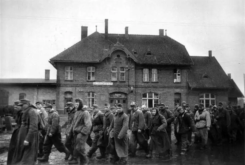 A column of German POWs in mixed winter uniforms marching past a brick railway station building in Landsberg, East Prussia, photographed by Emmanuil Evzerikhin.