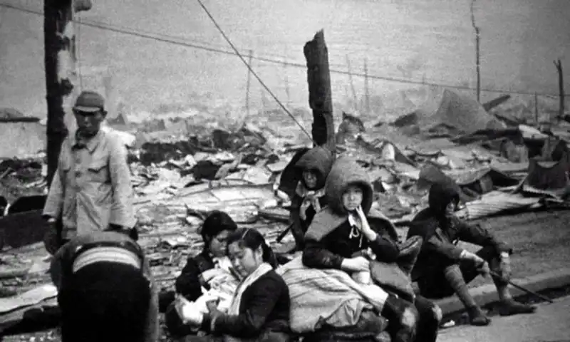 Group of displaced Japanese civilians, including women with young children, sitting on a curb surrounded by destroyed buildings in Tokyo, photographed by Kōyō Ishikawa in March 1945.