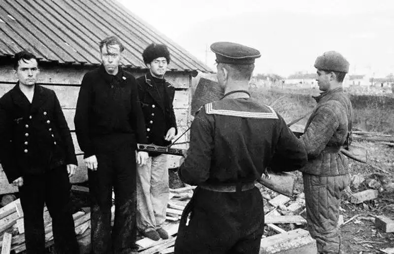 Black and white archival photo showing a Soviet sailor with a PPSh submachine gun guarding three captured German Kriegsmarine sailors in black uniforms near a wooden building, spring 1944