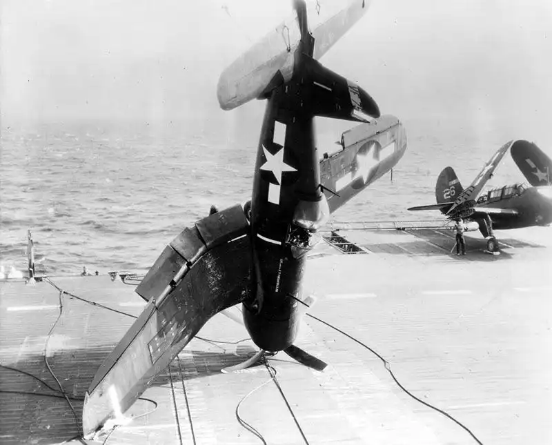 US Navy F4U Corsair fighter balancing vertically on its engine hub after landing arrester wire engagement on aircraft carrier deck.
