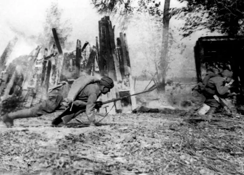 Black and white action photo of Soviet infantrymen advancing with bayoneted rifles past burning wooden houses in a village in 1943.