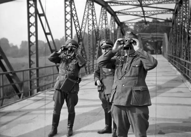 Black and white historical photo by Czesław Datka showing three German army officers in uniform looking through binoculars on a steel bridge in Bohumín, 1938.
