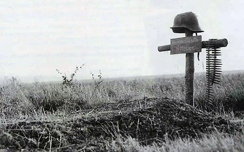 Black and white historical photo showing a rough wooden cross on a German field grave in a field, adorned with a dented steel helmet and a machine gun belt, autumn 1943.