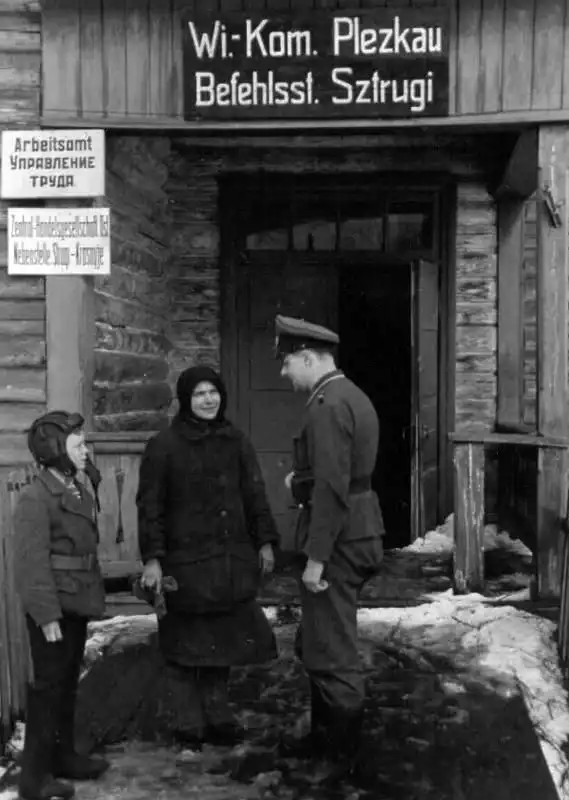 Young translator Kostya Slonov wearing a Red Army tank helmet talking to a Wehrmacht NCO outside the Arbeitsamt labor office sign in occupied Strugi Krasnye 1943.