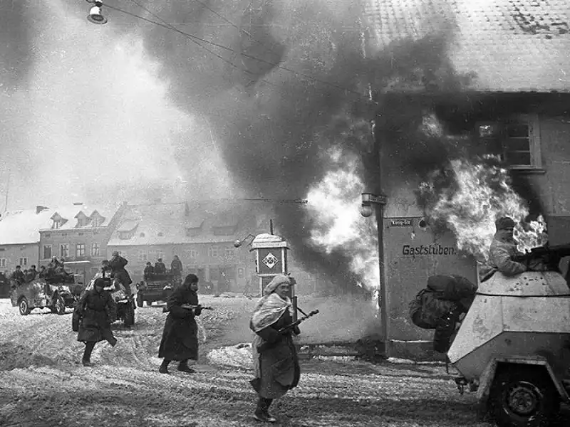 Red Army soldiers advance through snow past a burning German building alongside a BA-64 armored car and M3A1 Scout Cars in Mühlhausen, East Prussia.