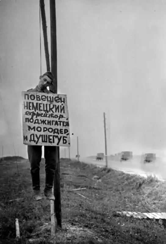 Historical archival photo of an executed German soldier hanging from a roadside pole in Belorussia in 1944 with a handwritten warning sign attached to his uniform.