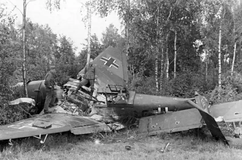 Black and white WWII archival photo of a burnt-out German Ju 87 Stuka dive bomber wreckage surrounded by birch trees in a Soviet forest in 1941.