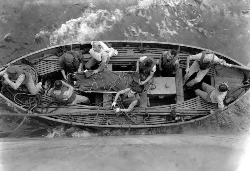 Black and white overhead photograph showing US Navy sailors in a small rescue boat surrounding a stretcher with a wounded crew member in the Pacific Ocean, 1945.