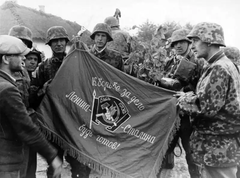 Waffen-SS soldiers in camouflage smocks holding a red Soviet Young Pioneer flag while a local resident attempts to translate the inscription in July 1941.