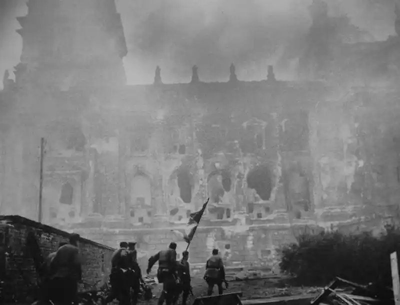 A Soviet assault group with the Red Banner advances toward the heavily damaged Reichstag under heavy fire.