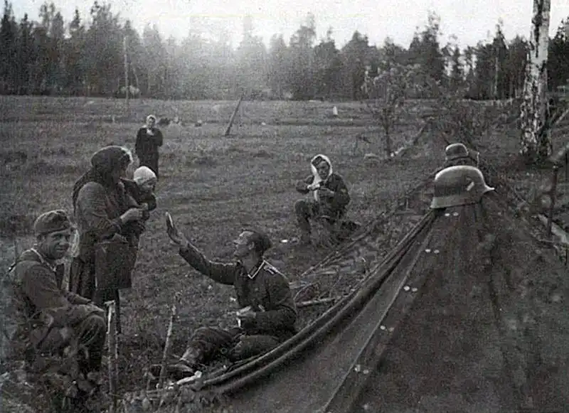 Black and white historical photo by Georg Gundlach showing a seated German soldier handing a piece of bread to a woman holding a young child near a canvas tent in 1942.