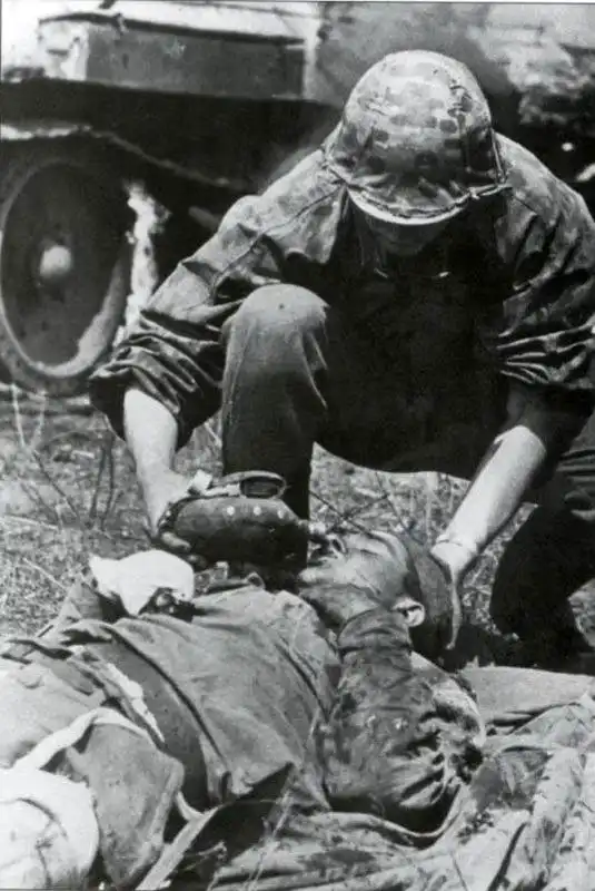Waffen-SS soldier in camouflage smock holding a field canteen to the lips of a wounded Soviet tanker lying near a damaged T-34 tank in July 1943.